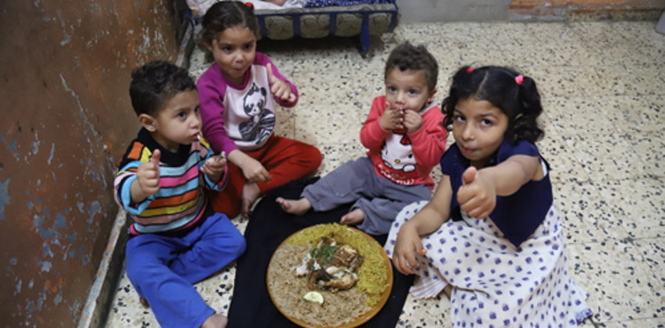 Four children sitting on the floor sharing a meal, with a baby sleeping in the background.