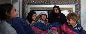 Sadaqah jariyah for parents A group of children holding white bags with the 'Humanitarian Aid' logo, standing in front of a banner that reads 'British International Waqf.' In the background, there's a mural depicting two children in school uniforms and a tree, suggesting the setting might be a classroom or school environment. The image reflects a charitable event where children are receiving aid or gifts.