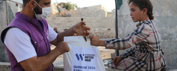Hadith about benefits of sadaqah A humanitarian worker wearing a mask and a purple vest hands a Qurbani 2024 Syria aid bag from International British Waqf to a young girl sitting on a stone wall in a rural area.
