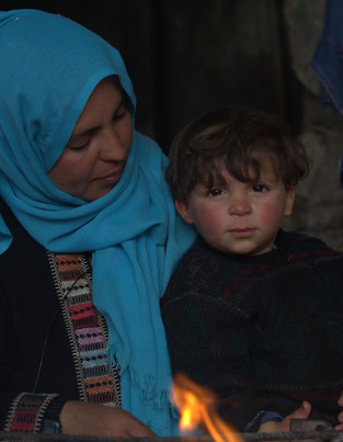 Is Giving Money To Parents Sadaqah A woman wearing a blue headscarf and a black garment with colorful embroidery is sitting beside a child who is wearing a dark sweater. There is a small fire burning in front of them. In the background, there is a wooden shelf with various items, including a television and some clothes.