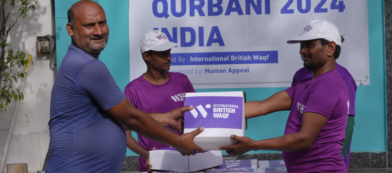 How To Do Sadaqah For Someone Else A man in a blue shirt receives a Qurbani 2024 food box from a volunteer in a purple shirt and white cap, during a distribution event in India organized by International British Waqf and implemented by Human Appeal.