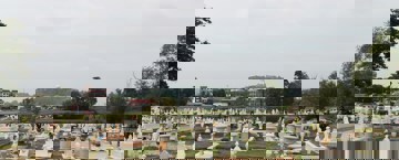 Best Sadaqah Jariyah For Deceased A wide view of a Muslim cemetery with neatly arranged graves, most of them white with decorative headstones. In the background, there are trees, buildings, and a hill with a prominent white structure on top.