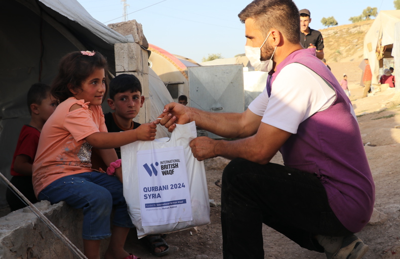 How To Do Sadaqah For Someone Else A humanitarian worker wearing a mask and purple vest hands a Qurbani 2024 food bag labeled "International British Waqf" to a young girl in a refugee camp in Syria, as two boys sit beside her.