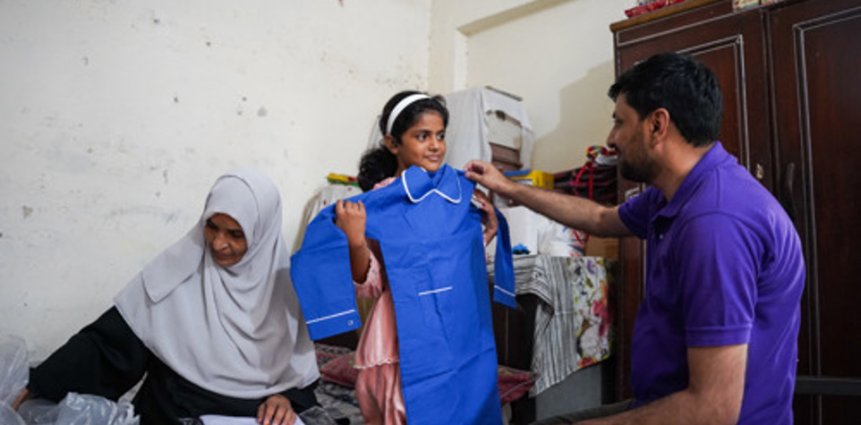 A young girl holds up a blue school uniform while a man and a woman sit beside her.