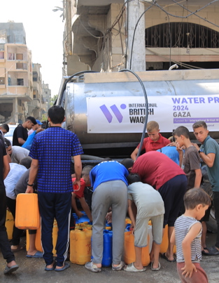 what is the meaning of waqf A group of people, including children and young men, gather around a water tanker in a damaged area, filling plastic containers with water. The banner on the tanker indicates a 2024 water project in Gaza, funded by "International British Waqf".